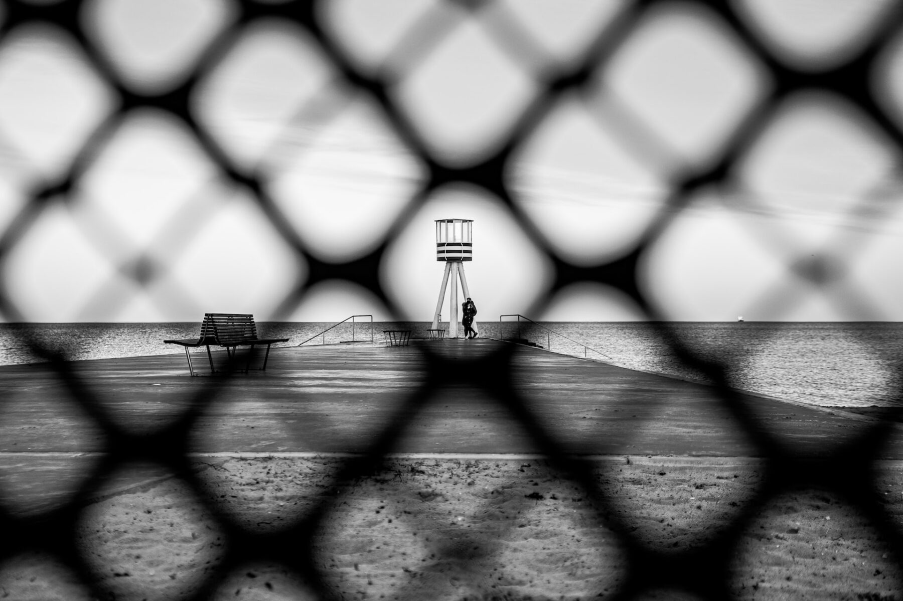 Tower and couple in background at the end of a pier with a fence in the foreground. Photo by Marco Chilese on Unsplash